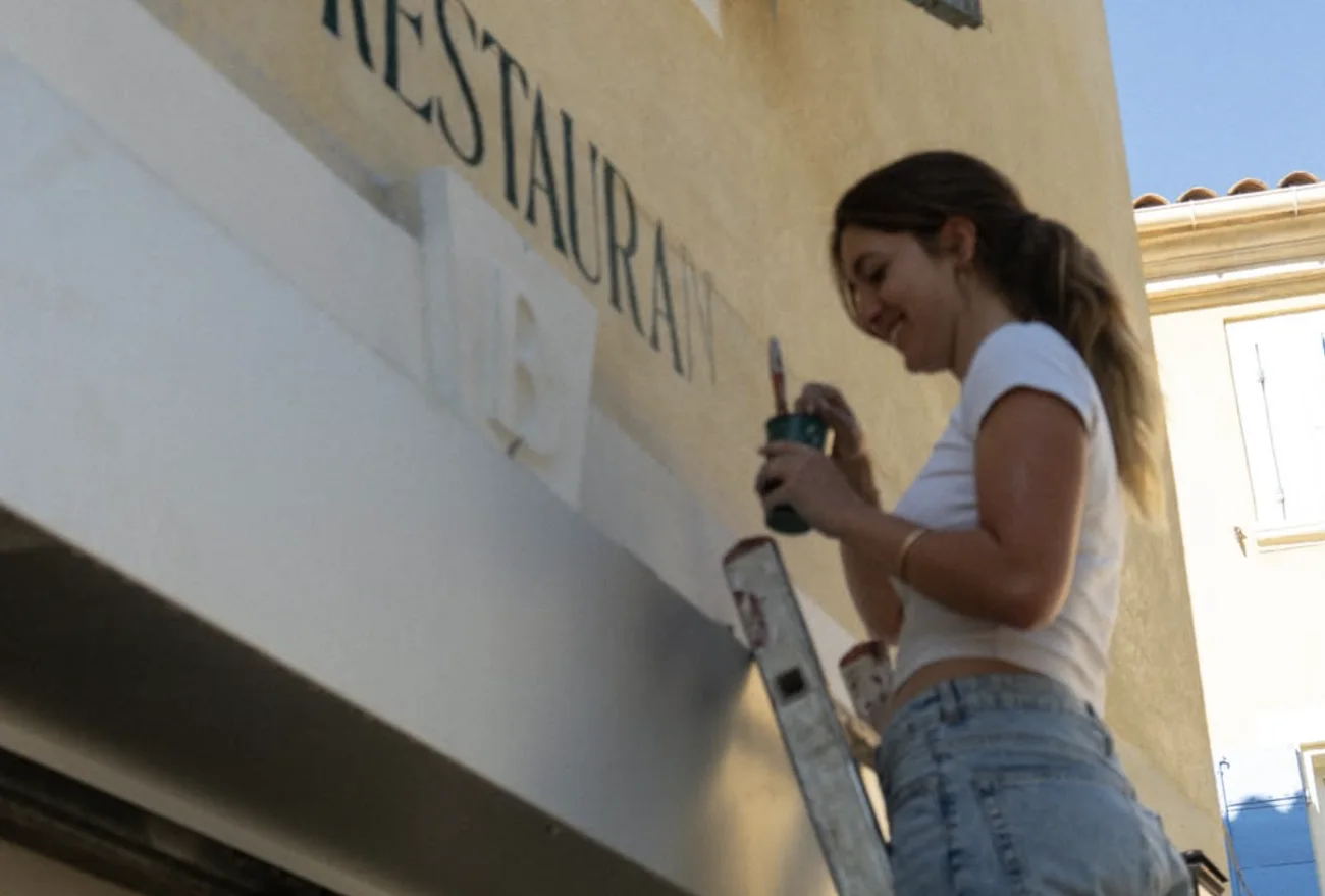 Iona sourit, pinceau et pot de peinture à la main, sur l'échelle